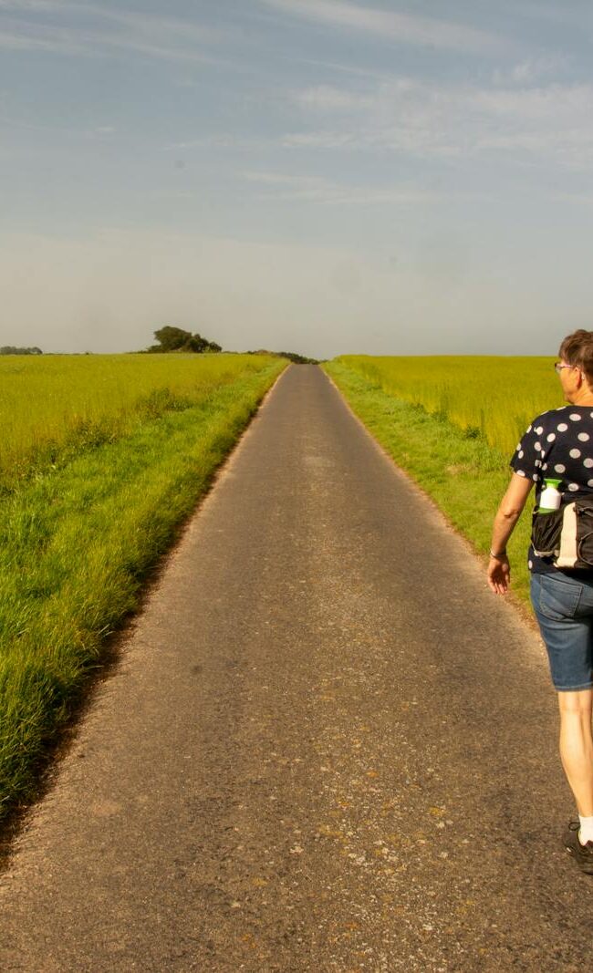 A woman walks on a road between lush green fields under a clear sky.