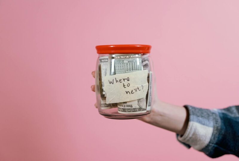 A hand holding a glass jar filled with dollar bills labeled 'Where to next?' against a pink background, symbolizing savings and future planning.