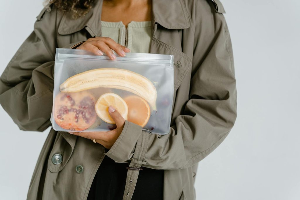 Person holding a transparent eco-friendly bag filled with fresh fruits including banana and orange.