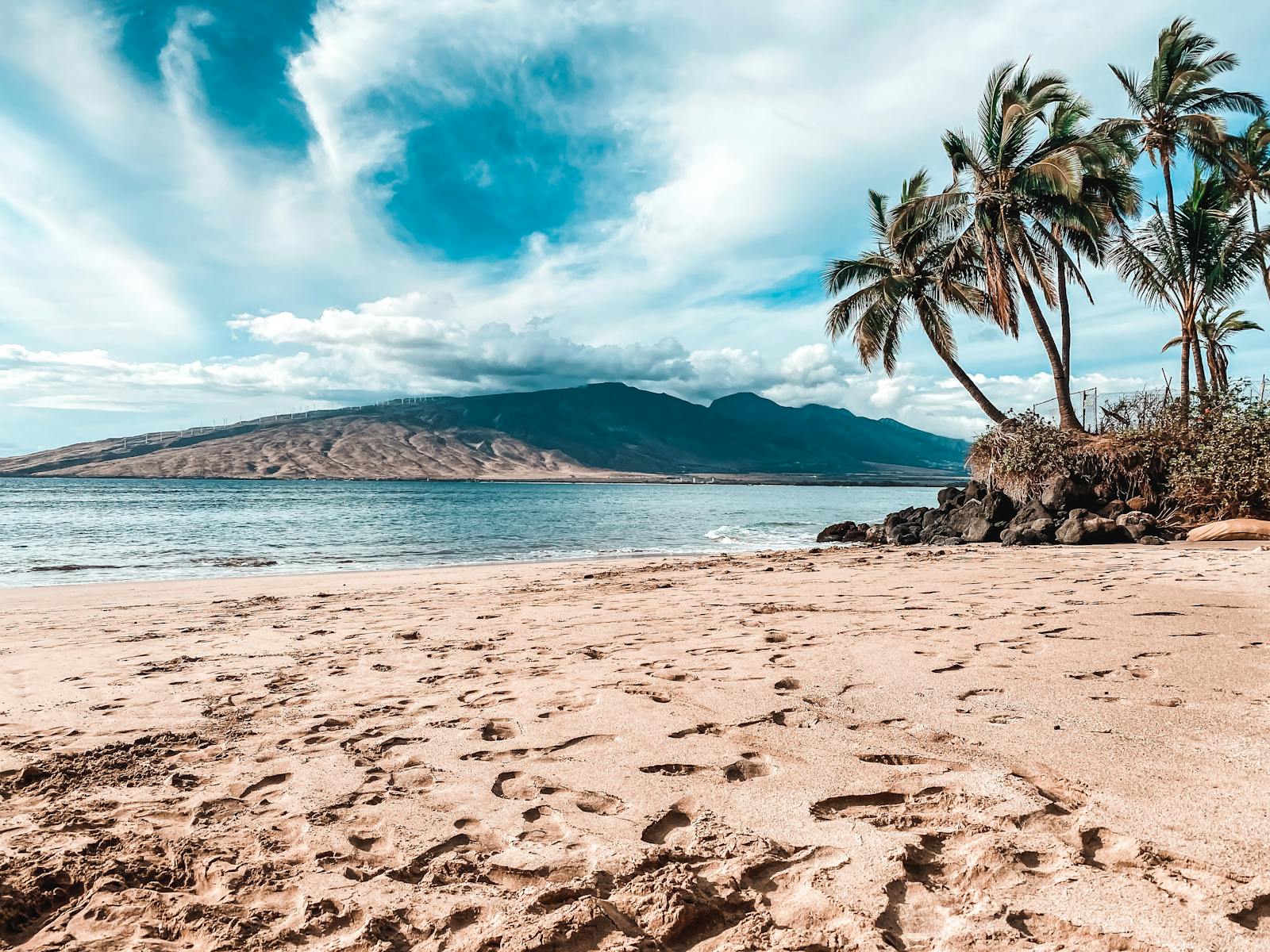 Idyllic tropical beach with palm trees, footprints in the sand, and a distant mountain view.