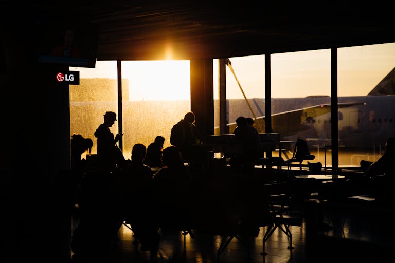 Travelers sitting in an airport terminal at sunset, silhouetted against the backdrop of an airplane.